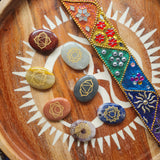 Set of seven chakra stones engraved with gold symbols, arranged on a wooden tray beside a colorful beaded decoration