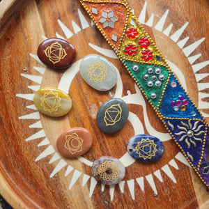 Set of seven chakra stones engraved with gold symbols, arranged on a wooden tray beside a colorful beaded decoration