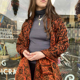 Person wearing a patterned orange and black coat in front of a store window.